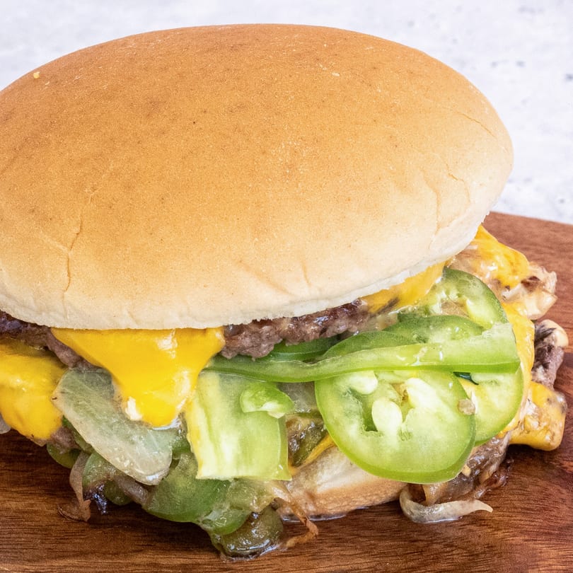 A Campus Burgers cheeseburger with melted cheese, green peppers, and onions on a wooden surface.