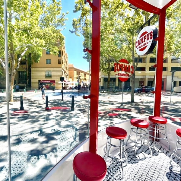 A Campus Burgers outdoor counter with red stools and a view of a sunny street.