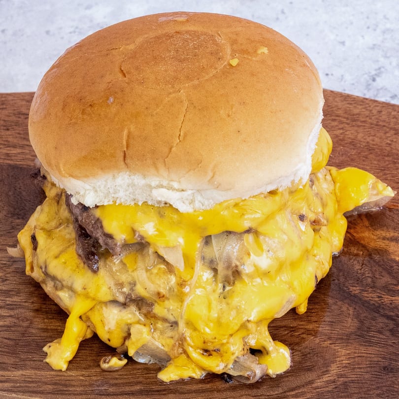 A Campus Burgers cheeseburger, with melted cheese and grilled onions spilling onto a wooden board.