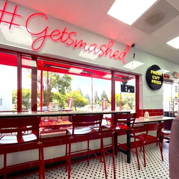 Interior of a restaurant with red bar stools, white and red tiled walls, and a neon "#Getsmashed" sign.